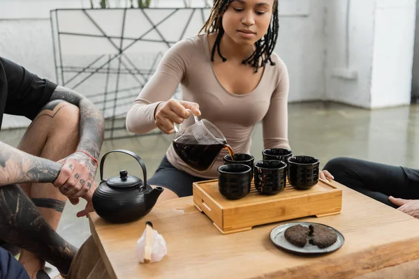 Mujer afroamericana con rastas vertiendo puré en tazas de té tradicionales cerca del hombre tatuado - foto de stock