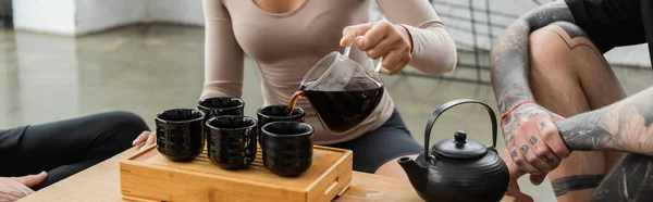 Vue recadrée de femme afro-américaine versant puer dans des tasses à thé traditionnelles près de l'homme tatoué, bannière — Photo de stock