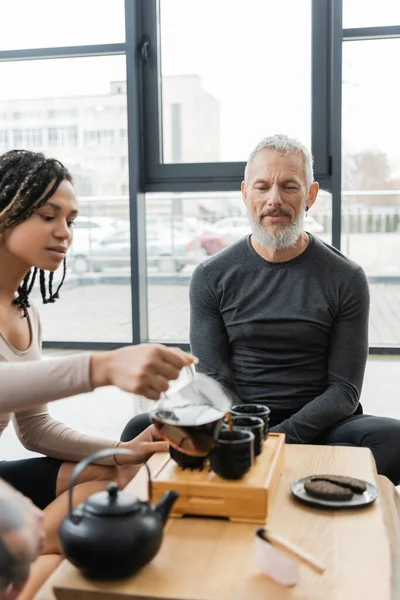 African american woman with dreadlocks pouring puer into traditional tea cups near middle aged man — Stock Photo