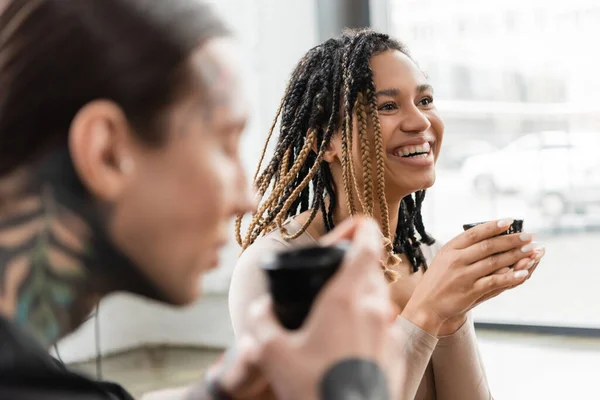 Весела афроамериканська жінка з дредами, що тримають япанську чашку з чистішим чаєм біля татуйованого чоловіка на розмитому передньому плані. — стокове фото