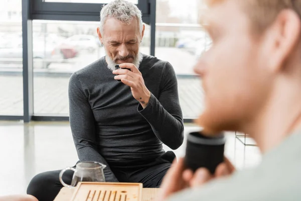 Bearded middle aged man smelling puer tea near bearded friend on blurred foreground — Stock Photo