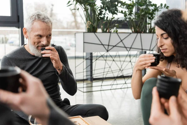 Happy mature man drinking puer tea while looking at middle eastern woman in yoga studio — Stock Photo