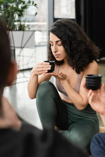 Curly middle eastern woman smelling puer tea near people in yoga studio — Stock Photo