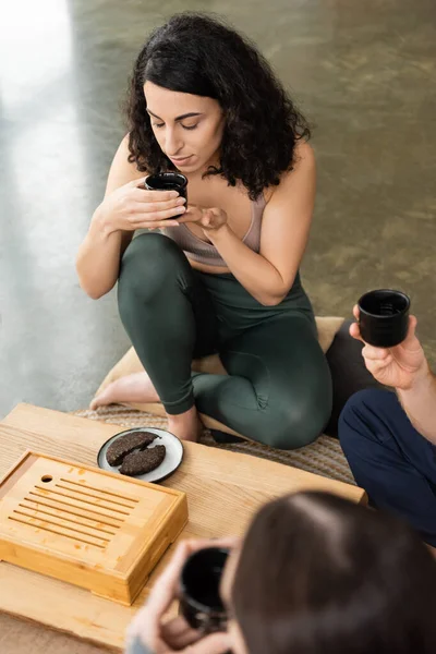 Curly middle eastern woman smelling puer tea near men in yoga studio — Stock Photo