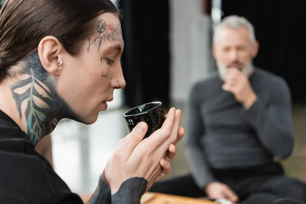 Vista lateral del hombre tatuado sosteniendo la taza china con té puer elaborado cerca del hombre de mediana edad sobre un fondo borroso - foto de stock