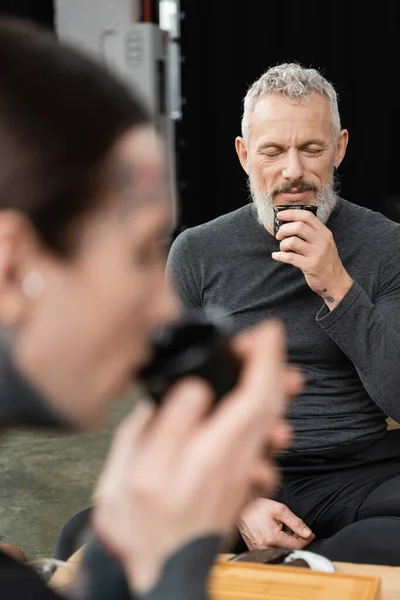 Homem de meia idade barbudo com olhos fechados cheirando chá puer perto homem tatuado em primeiro plano borrado — Fotografia de Stock