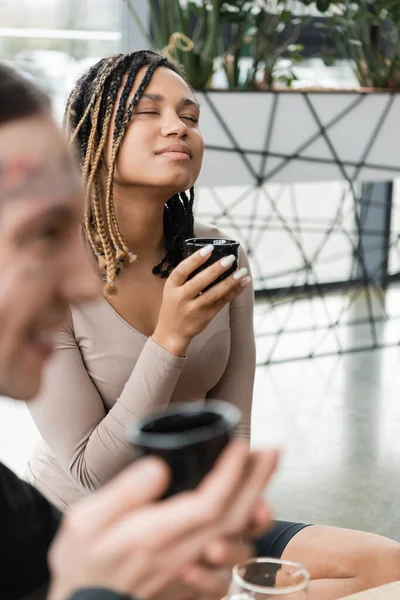 Heureux afro-américain femme avec dreadlocks tenant tasse avec puer thé près de tatoué homme sur flou premier plan — Photo de stock