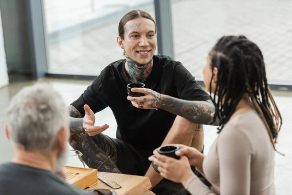 Happy and tattooed man holding Chinese tea cup and talking with african american woman in yoga studio — Stock Photo