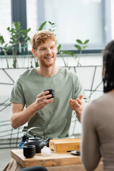 Happy man with red hair and beard holding Chinese cup and talking with african american woman — Stock Photo
