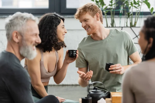 Happy man with red hair holding Chinese cup and talking with middle eastern woman near people on blurred foreground — Stock Photo