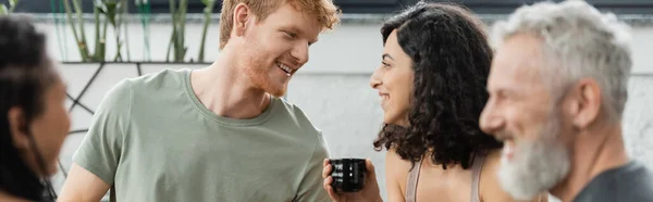 Happy man with red hair talking with middle eastern woman with chinese cup near people on blurred foreground, banner — Stock Photo