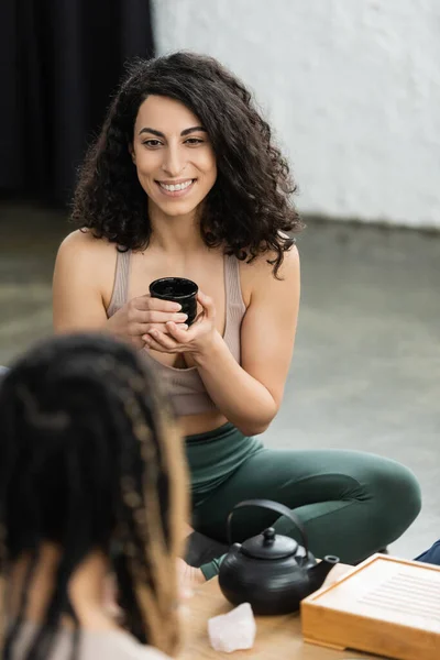 Happy middle eastern woman with curly hair holding Chinese cup during tea ceremony in yoga studio — Stock Photo