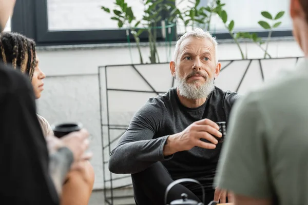 Bearded middle aged man holding Chinese cup with puer tea near people in yoga studio — Stock Photo