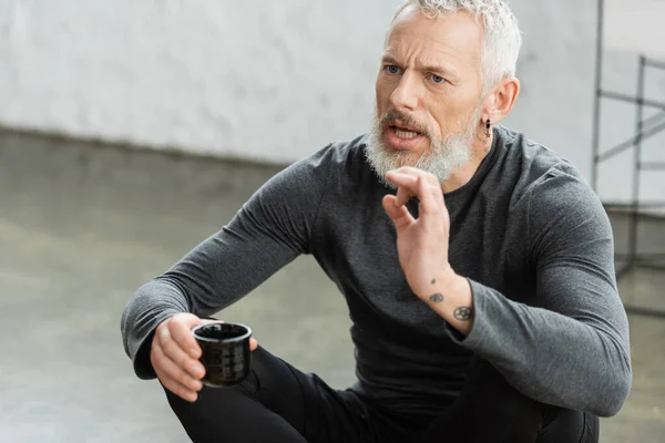 Bearded middle aged man with tattoos holding Chinese cup with puer tea while talking in yoga studio — Stock Photo