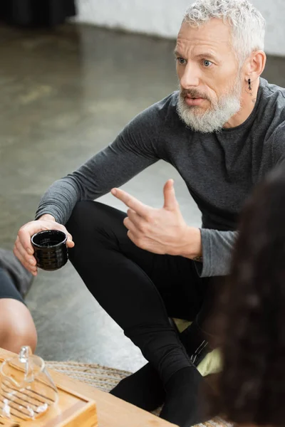 Middle aged man with tattoos holding Chinese cup with puer tea while talking in yoga studio — Stock Photo