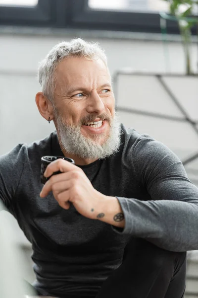 Positive middle aged man with tattoos holding Chinese cup with puer tea in yoga studio — Stock Photo