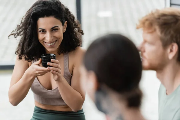 Joyful middle eastern woman with curly hair smiling while drinking puer tea near men on blurred foreground — Stock Photo