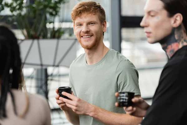 Cheerful man with red hair holding japanese cup with puer tea near people in yoga studio — Stock Photo