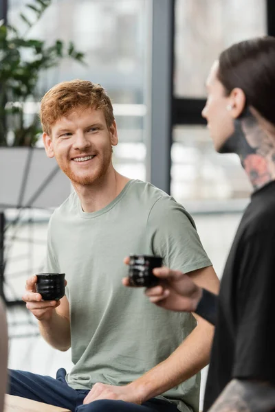 Positive man with red hair holding Japanese cup with puer tea near tattooed friend in yoga studio — Stock Photo