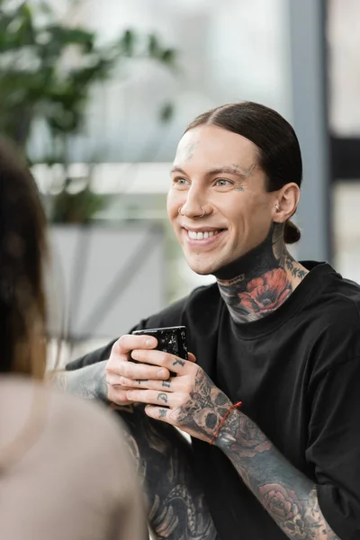 Homme heureux et tatoué souriant tout en tenant une tasse japonaise avec du thé près de la femme sur le premier plan flou — Photo de stock