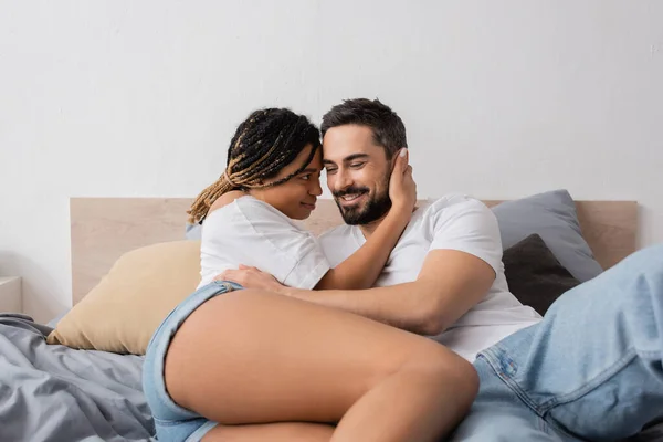 Sexy afrikanisch-amerikanische Frau und fröhliche bärtige Mann entspannt und umarmt auf dem Bett zu Hause — Stockfoto