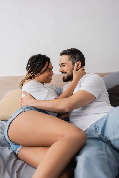 Casal interracial satisfeito em camisetas brancas olhando um para o outro e abraçando na cama em casa — Fotografia de Stock