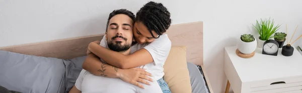 Vista ad alto angolo della donna afroamericana tatuata che abbraccia l'uomo barbuto sdraiato con gli occhi chiusi sul letto a casa, striscione — Foto stock