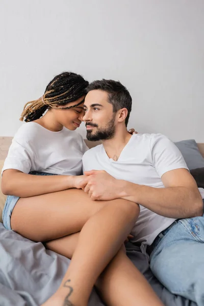 Barbudo hombre cogido de la mano de bastante africana americana novia mientras descansa en el dormitorio en casa - foto de stock