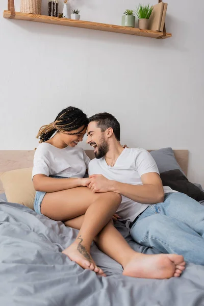 Excité homme barbu riant près pieds nus afro-américaine femme couchée sur le lit à la maison — Photo de stock