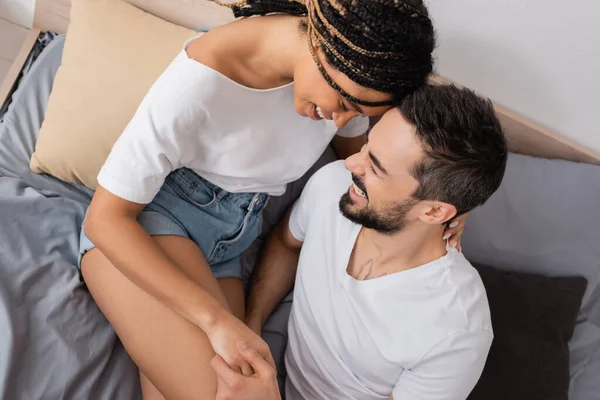 Top view of carefree interracial couple in white t-shirts holding hands and smiling on bed at home — Stock Photo