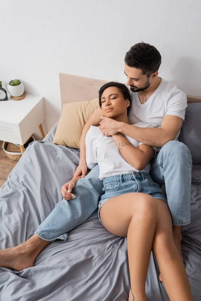 High angle view of bearded man embracing african american girlfriend lying in white t-shirt and denim shorts in bedroom — Stock Photo
