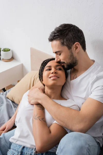 Uomo barbuto abbracciare giovane e felice donna africana americana sorridente sul letto a casa — Foto stock