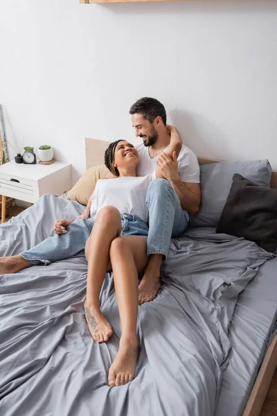 Fröhliches multiethnisches Paar, das es sich zu Hause auf dem Bett gemütlich macht und einander anlächelt — Stockfoto