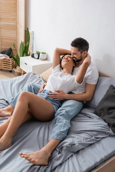 Joven mujer afroamericana en pantalones cortos de mezclilla abrazando al hombre barbudo mientras se relaja en la cama en casa — Stock Photo