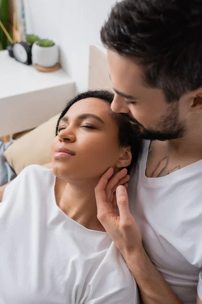 High angle view of bearded man touching young african american woman relaxing with closed eyes in bedroom at home — Stock Photo