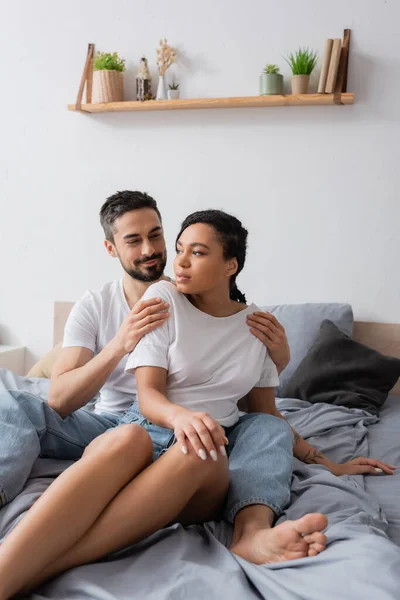 Smiling man hugging african american girlfriend in white t-shirt looking away on bed at home — Stock Photo