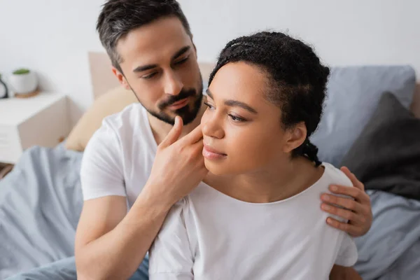 Bearded man touching face of charming and dreamy african american woman looking away in bedroom at home — Stock Photo