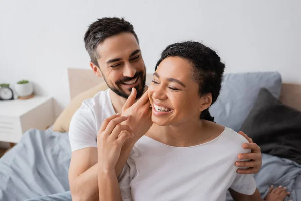 Homme barbu souriant touchant visage de femme afro-américaine ravie dans la chambre à coucher à la maison — Photo de stock
