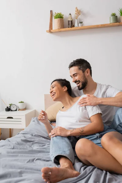 Excité homme barbu étreignant femme américaine africaine gaie tout en s'amusant sur le lit à la maison — Photo de stock