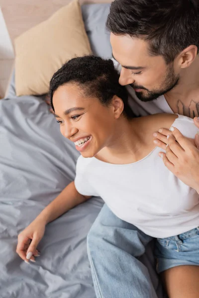 Vue grand angle de femme afro-américaine gaie souriant près barbu tatoué homme dans la chambre à coucher à la maison — Photo de stock