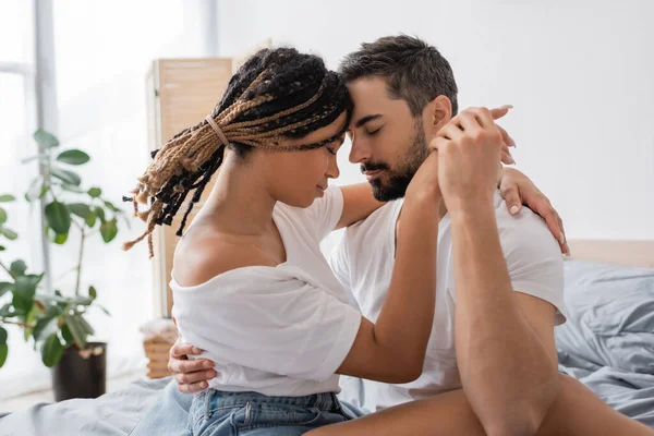 Seitenansicht eines bärtigen Mannes und einer afrikanisch-amerikanischen Frau mit Dreadlocks, die Händchen halten und sich mit geschlossenen Augen im Schlafzimmer umarmen — Stockfoto