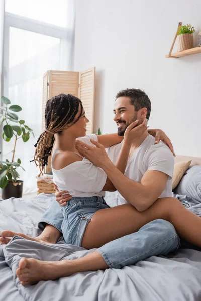 Fröhliche interrassische Paar in weißen T-Shirts umarmen und lächeln einander an, während sie auf dem Bett zu Hause sitzen — Stockfoto