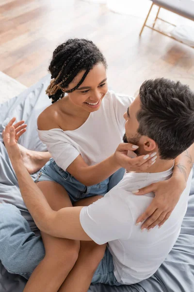 Vista ad alto angolo di felice donna africana americana in t-shirt bianca che abbraccia il fidanzato mentre è seduto sul letto a casa — Foto stock