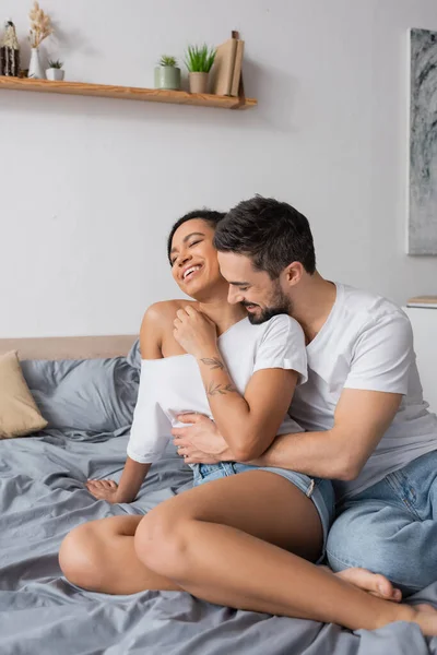 Bärtiger Mann im weißen T-Shirt umarmt unbeschwerte Afroamerikanerin, die zu Hause auf dem Bett sitzt — Stockfoto