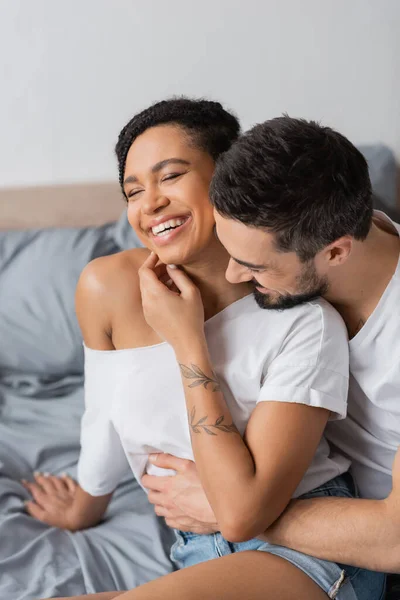 Homme barbu étreignant insouciante femme afro-américaine souriant avec les yeux fermés dans la chambre à coucher à la maison — Photo de stock