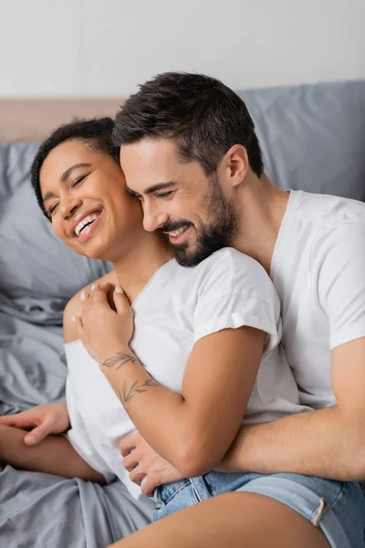 Hombre barbudo complacido abrazando a mujer afroamericana bonita sonriendo con los ojos cerrados en el dormitorio en casa - foto de stock
