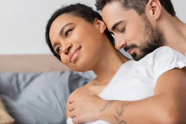 Улыбающаяся и мечтательная африканская американка, глядя в сторону бородатого мужчины, обнимающего ее в спальне дома — стоковое фото