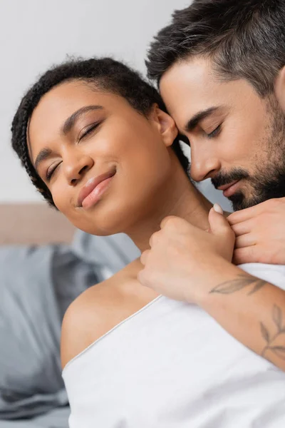 Encantadora mujer afroamericana sonriendo con los ojos cerrados mientras hombre barbudo abrazándola en el dormitorio en casa — Stock Photo