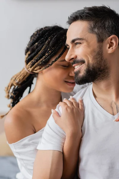 Jeune femme afro-américaine avec dreadlocks étreignant heureux homme barbu regardant loin à la maison — Photo de stock