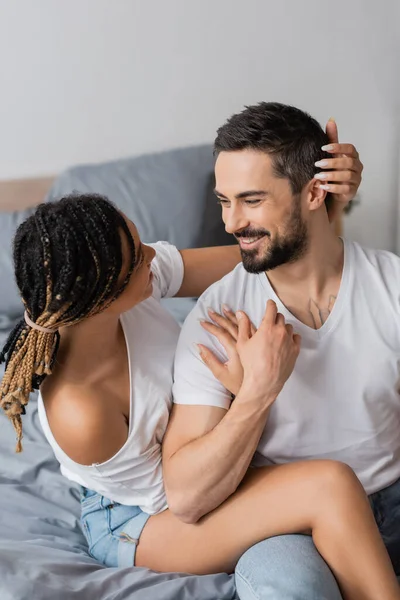 Carefree bearded man and african american woman with dreadlocks looking at each other on bed at home — Stock Photo